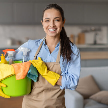 Confident young woman with beaming smile holding green bucket filled with colorful cleaning supplies and cloths, wearing yellow gloves and beige apron
