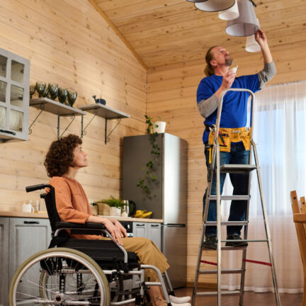 Young housewife with disability sitting in wheelchair in the middle of kitchen and looking at electrician changing bulbs in chandelier